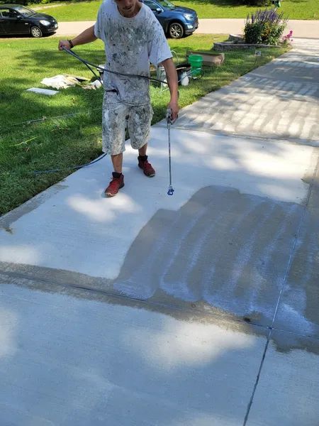 Person spraying concrete sealer on a driveway; wearing stained clothes, outside.