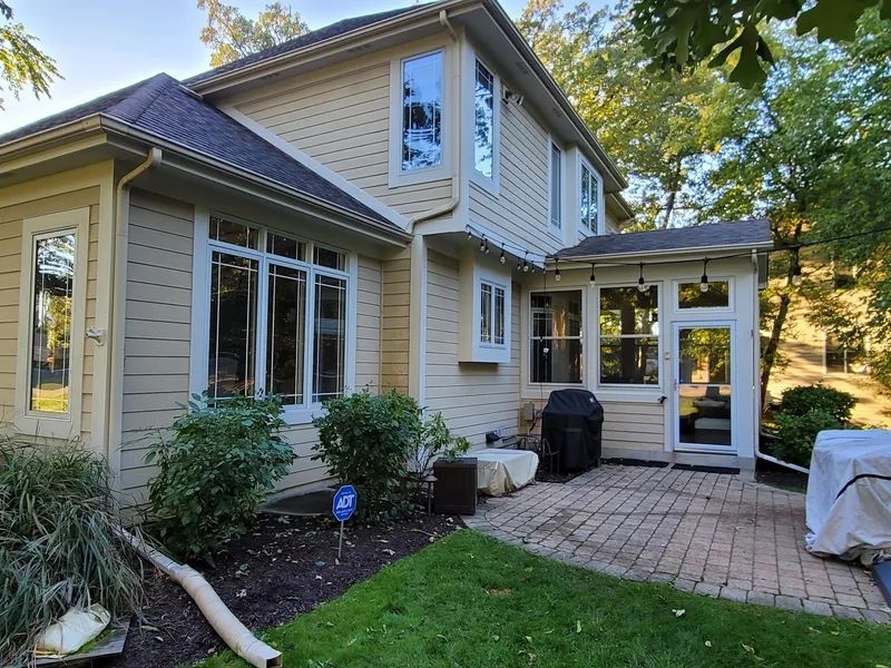 Two-story beige house with multiple windows, a patio, and lush greenery.