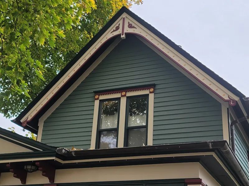 Green gabled roof with ornate trim and a double window. Part of a house, set against foliage.