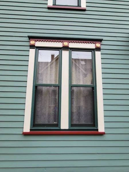 Double-hung window with ornate green trim, white accents, and red trim on a green clapboard house.