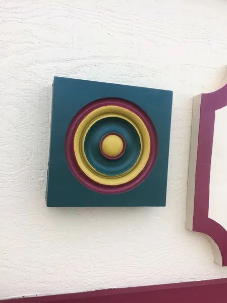 Square, teal wooden decoration with concentric rings of yellow, purple, and teal.