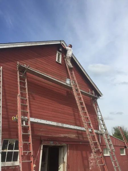 Person on ladder painting the red roof of a barn against a blue sky.