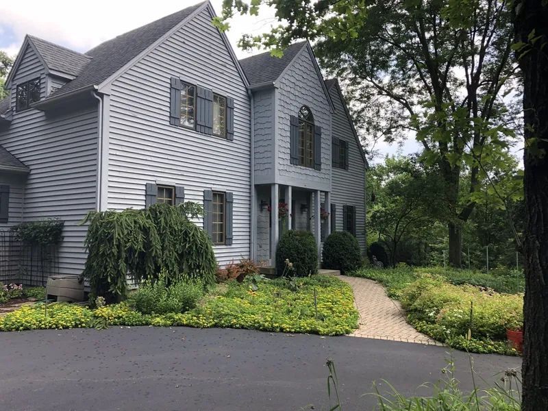 Two-story gray house with black shutters, a curved brick path, and lush landscaping.
