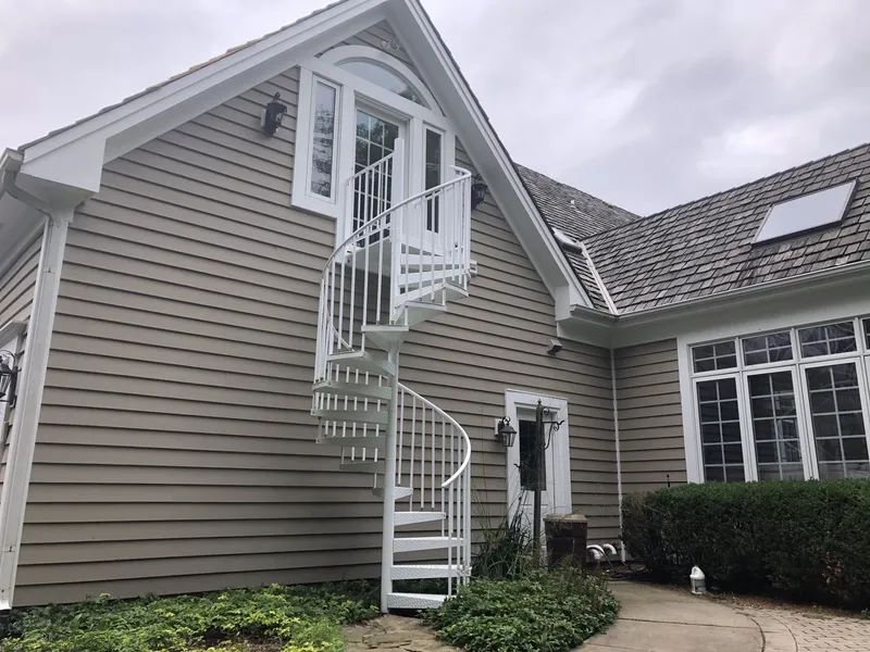 White spiral staircase against a two-story house with brown siding, leading to a white door.