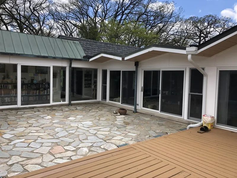 Patio with stone floor, deck, and white-framed sliding glass doors, surrounded by trees.