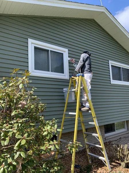 Person on a ladder painting window trim of a green-sided house. White trim and a blue sky are visible.