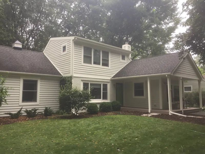 Tan house with dark roof, green lawn, and surrounding trees.
