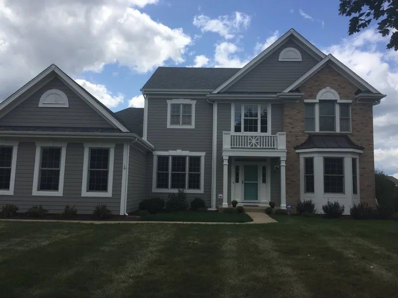 Two-story gray house with white trim, light blue door, and brick accent. Green lawn and cloudy sky.