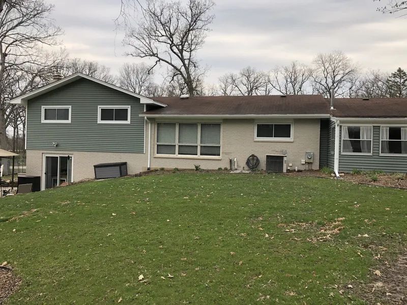 Back view of a house with green and beige siding on a grassy hill. Overcast sky, trees in the background.