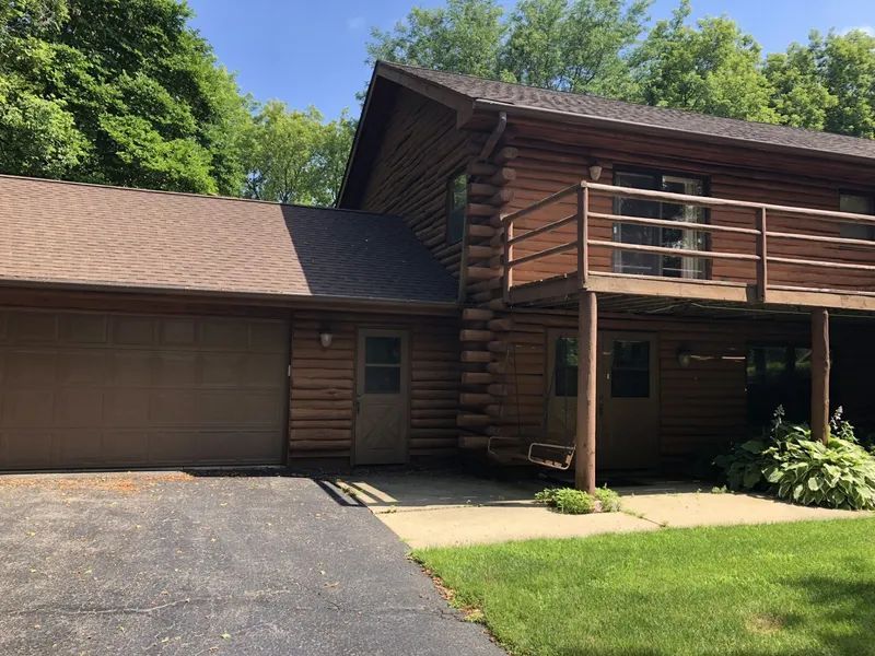 Log cabin with attached garage, brown siding, and a second-story deck. Driveway and green lawn.