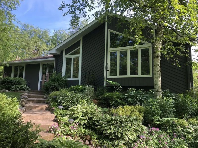Dark gray house with large windows, surrounded by lush green landscaping, stone steps leading up to the entrance.