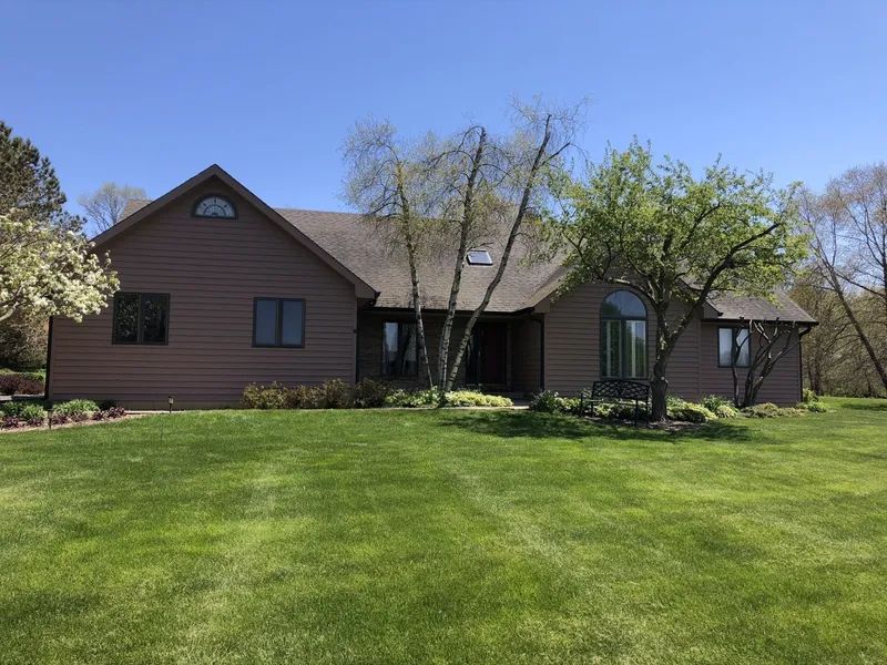 Brown house with arched window, tall trees, and green lawn under a clear blue sky.