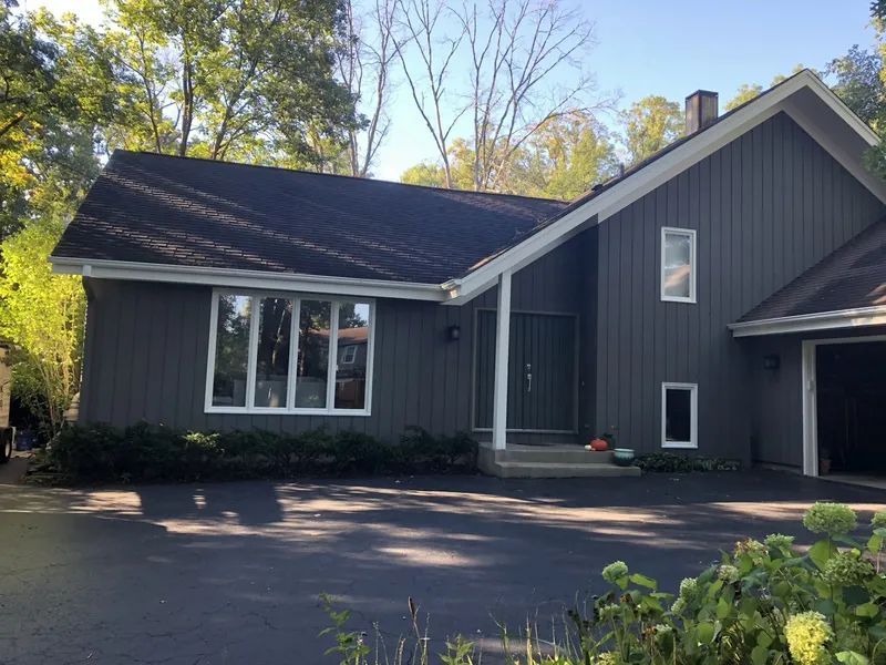 Gray house with dark roof and white trim, set in a driveway with trees in the background.