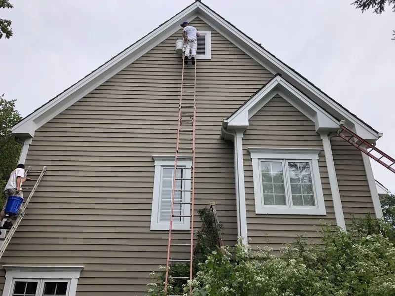 Two painters on ladders painting a two-story beige house with white trim under a cloudy sky.