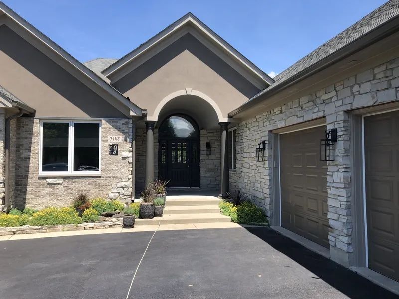 Stone-clad home with gray trim, arched doorway, black door, and two-car garage; asphalt driveway.