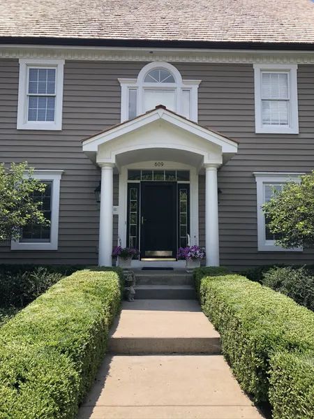 A two-story house with gray siding, white trim, and a concrete pathway lined with bushes.