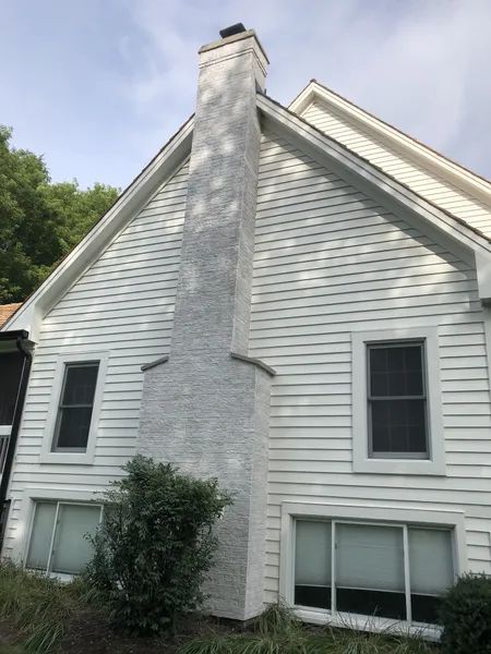 White house with a tall, textured chimney; two windows on the ground floor and two above, with green foliage at the base.