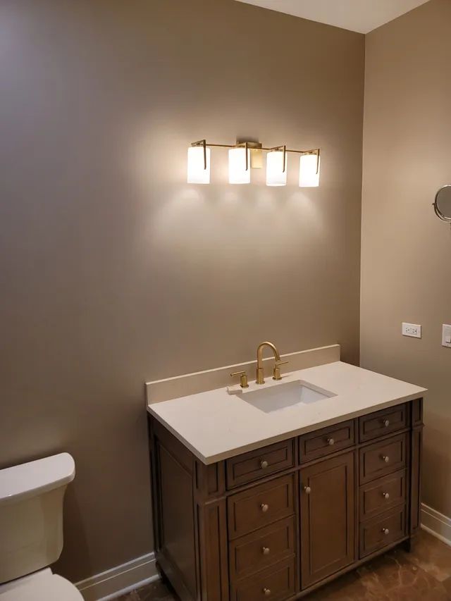 Bathroom with vanity, sink, toilet, gold fixtures, and light fixture against a tan wall.