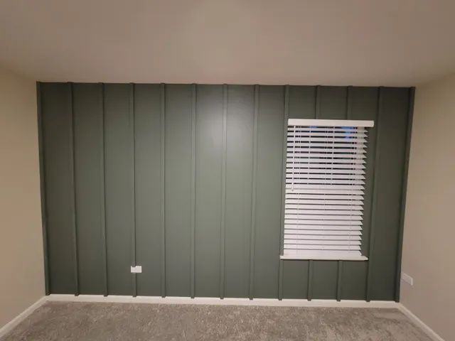 Green paneled accent wall with white blinds in a room with beige walls and a carpeted floor.