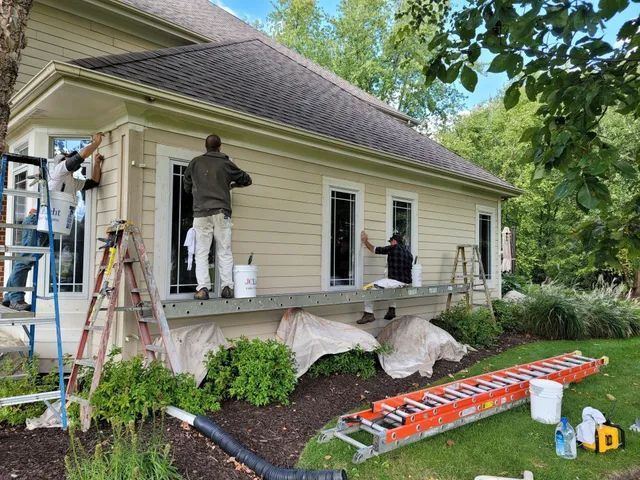 Three workers painting exterior of a light beige house, using ladders and scaffolding. Green grass and bushes surround.