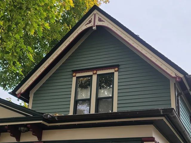 Gabled dormer with teal siding and decorative trim, set against a backdrop of green foliage.
