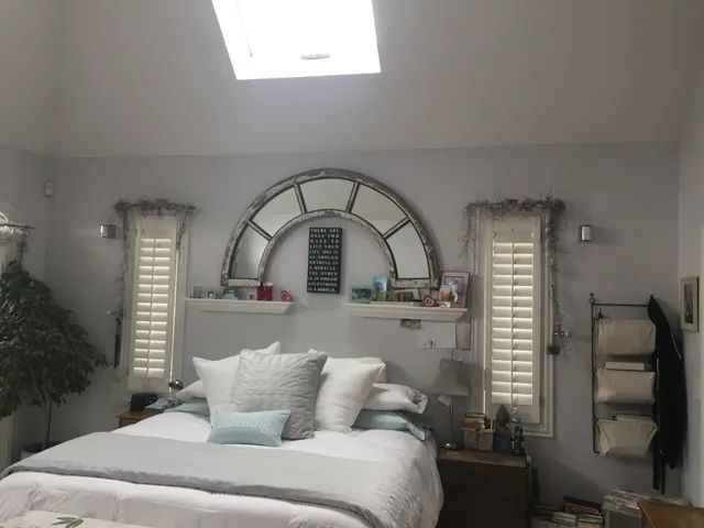Bedroom with white bed, large arched mirror, skylight, and shutters.