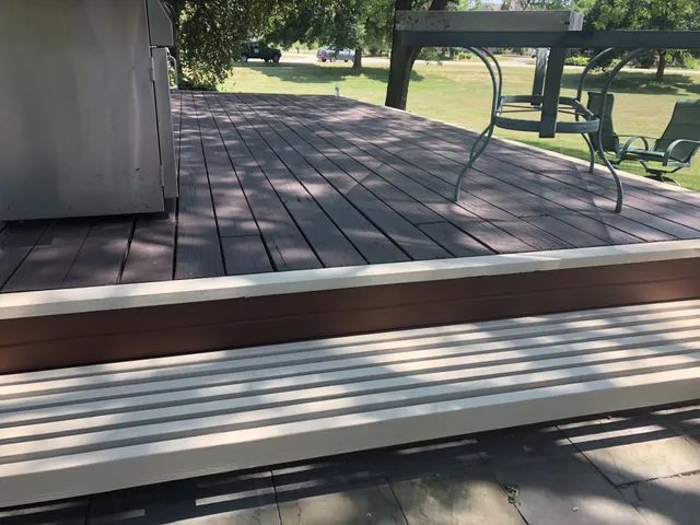 Wooden deck with brown and white composite boards, benches, and furniture in a park-like setting.