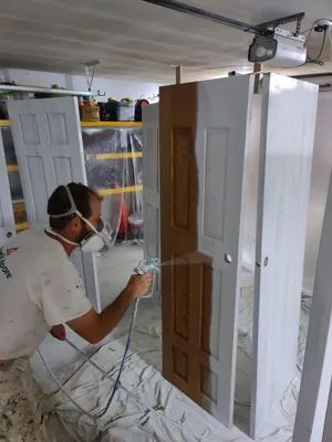 Man in respirator paints a brown door white with a spray gun in a garage.
