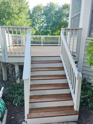 Wooden deck with stairs, light gray railing, and brown steps leading up. Green foliage surrounds it.