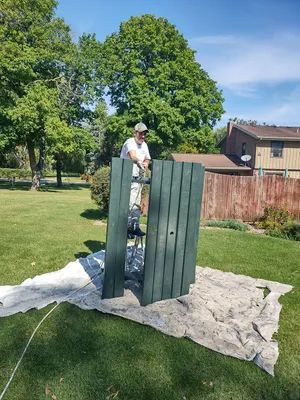 Man painting green wooden planks outdoors on a sunny day; green grass and trees in background.