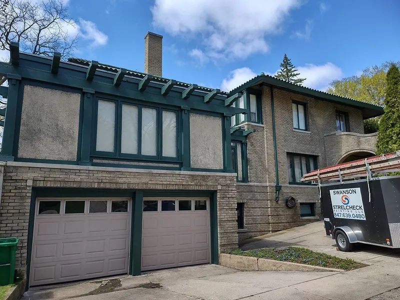 Two-story house with stone facade, green trim, and tan garage doors on a sunny day.