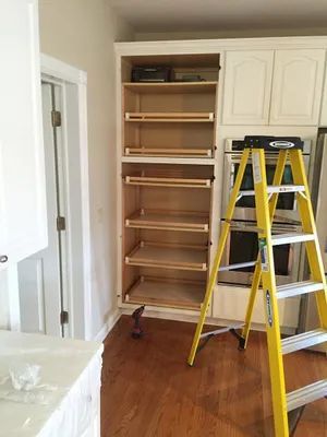 Tall white pantry with pull-out shelves is being installed. A yellow ladder leans against it in a kitchen with hardwood floors.