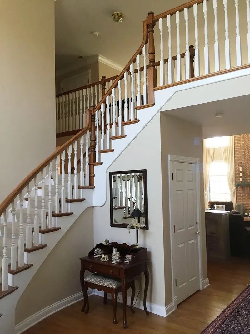 Wooden staircase with white balusters and brown handrails in a two-story foyer with beige walls and a small antique table.