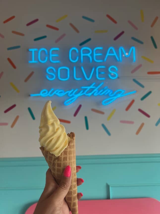Person holding a waffle cone with soft-serve ice cream. Neon sign says,