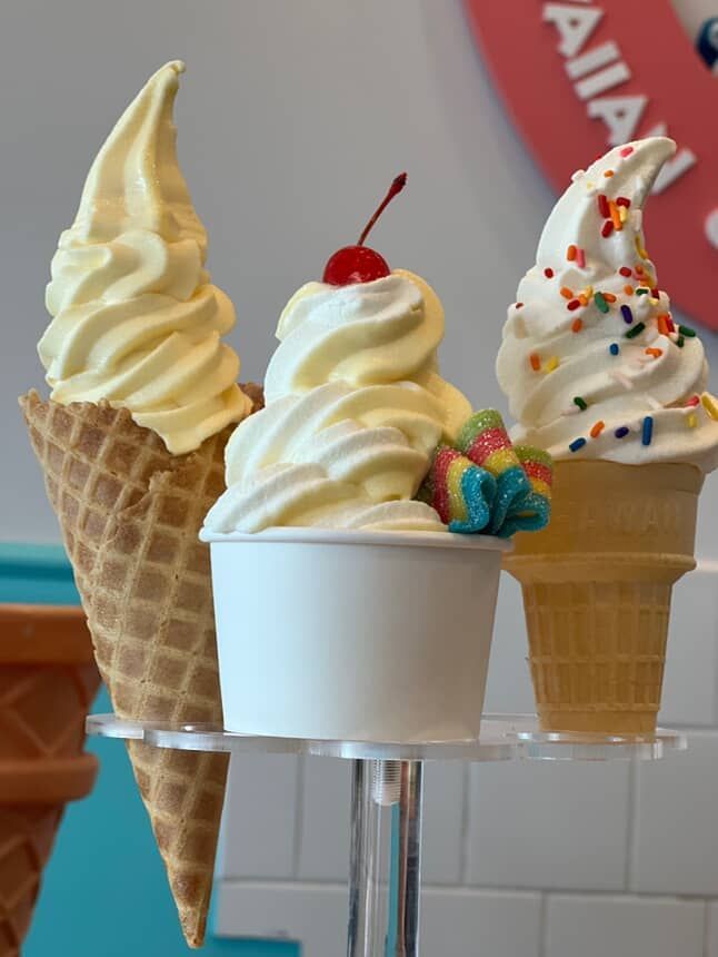 Three soft serve ice cream treats: cone, cup with cherry, and cone with sprinkles.