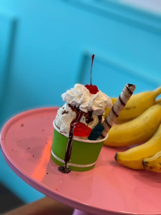 Ice cream sundae in a green cup, whipped cream, cherry, chocolate sauce, on pink plate with bananas.