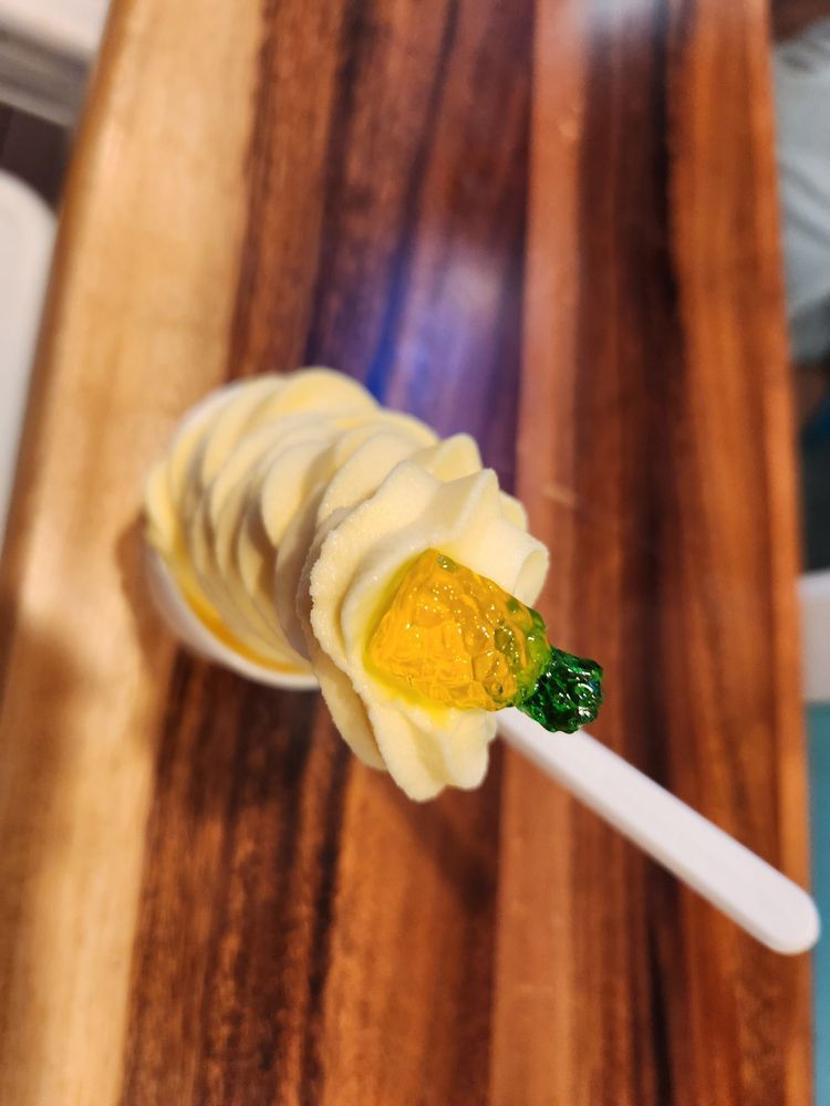 Soft-serve ice cream cone on a spoon, with a gummy pineapple at the top. On a wooden cutting board.