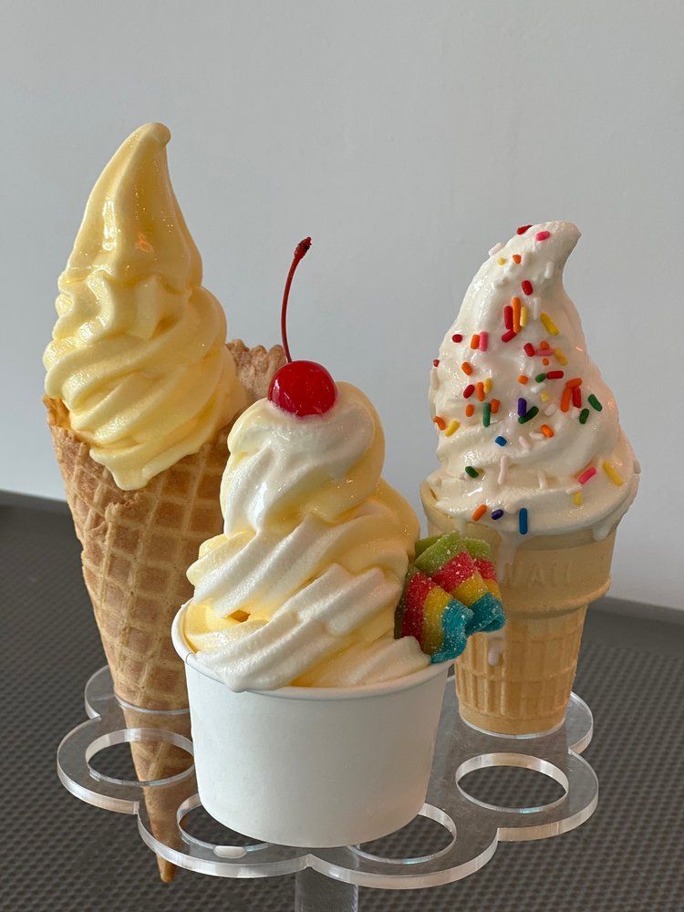 Three soft-serve ice cream cones: one lemon, one vanilla with sprinkles, and one swirl with a cherry, in a clear holder.