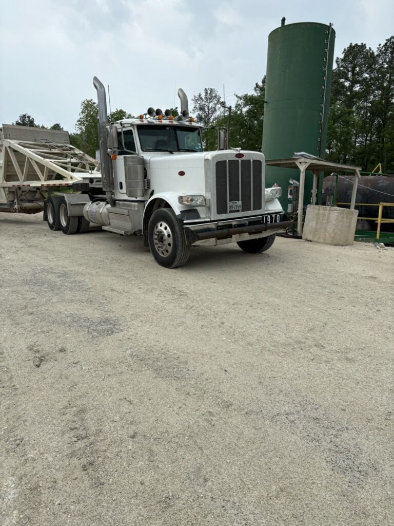 A white semi truck is parked in a gravel lot next to a green tank.