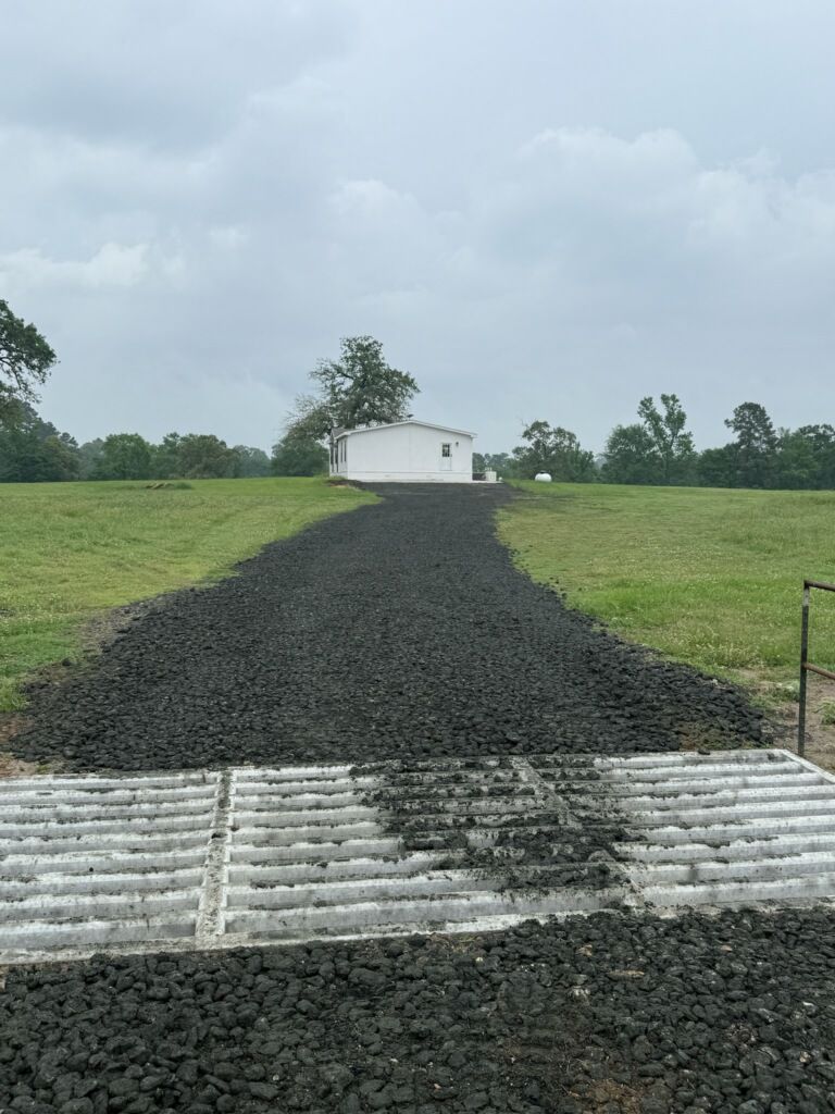A gravel road going through a grassy field with a white building in the background.