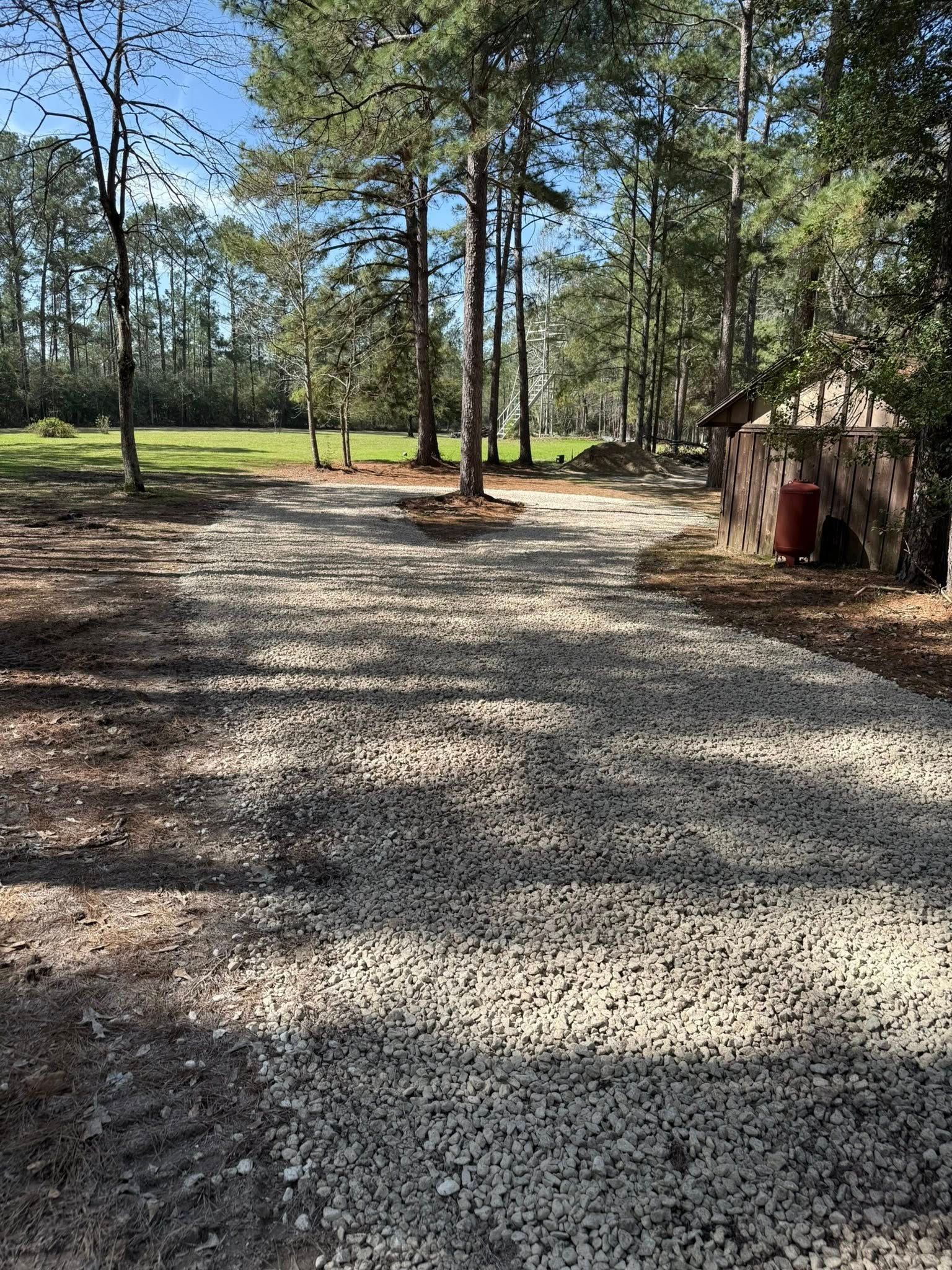 A gravel driveway leading to a field surrounded by trees on a sunny day.