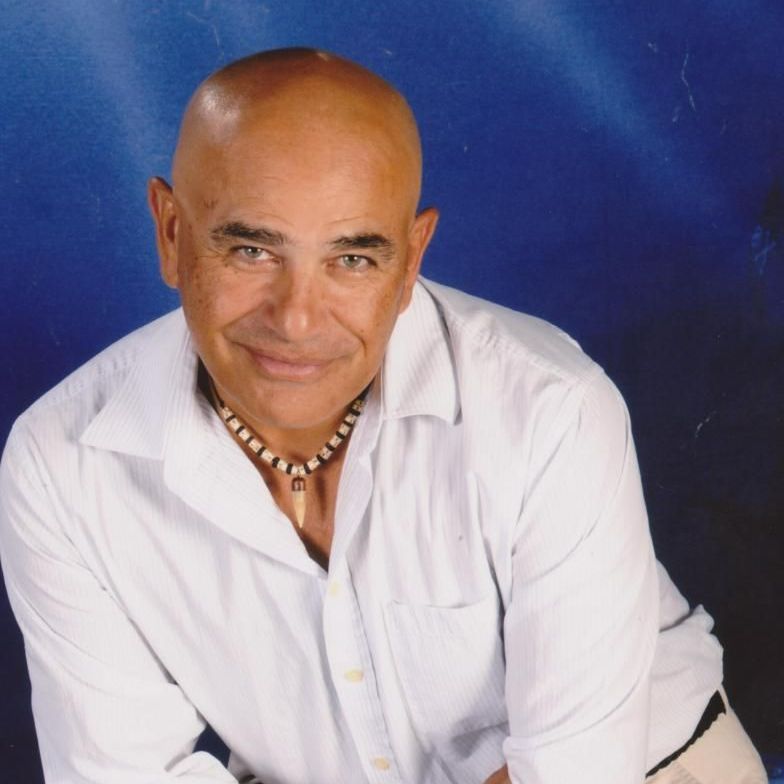 A bald man wearing a white shirt and a necklace smiles for the camera