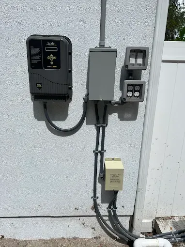 Wall-mounted electrical panel and outlet boxes beside a white exterior wall with conduit and cables.