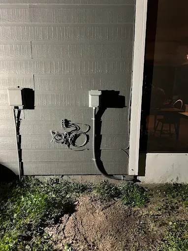 Exterior wall with two electrical boxes and a coiled cable above a dirt patch by a doorway at night