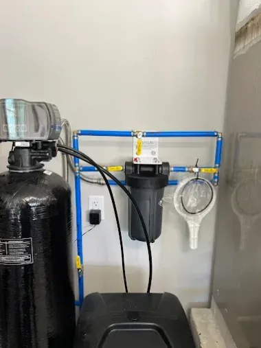 Water filtration setup with black tanks, blue piping, and a wall-mounted gauge in a utility room