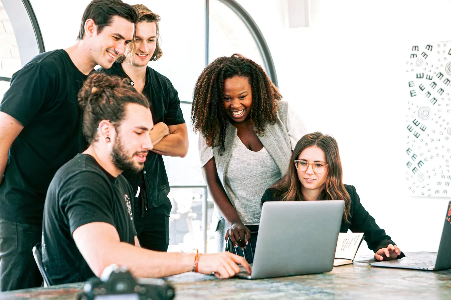 Five people look at a laptop screen together, smiling and collaborating at a table in a bright room.