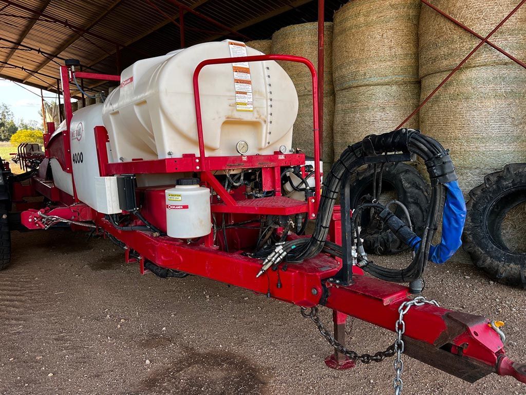 A red and white sprayer is parked in front of a pile of hay bales.