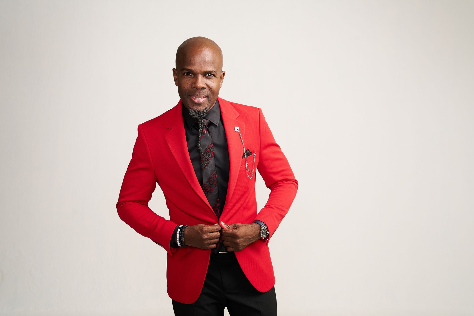 Man in a red blazer, black shirt and tie, holding lapels, smiling.