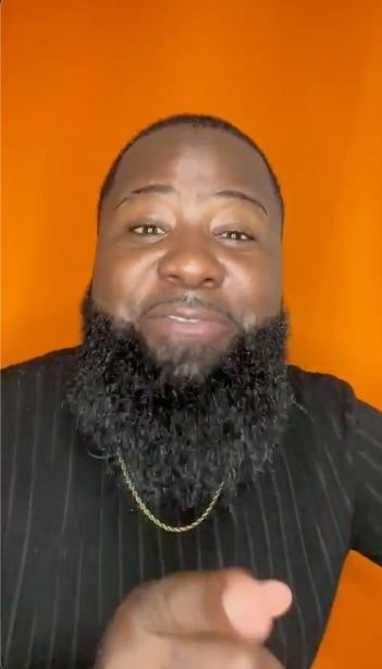 A man with a beard and a gold chain is pointing at the camera.