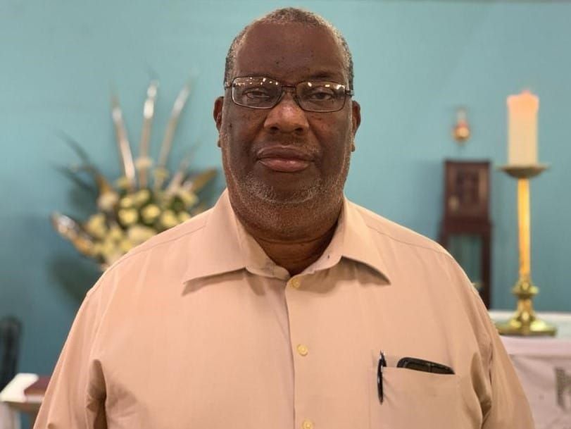 A man wearing glasses and a tan shirt is standing in front of a candle in a church.